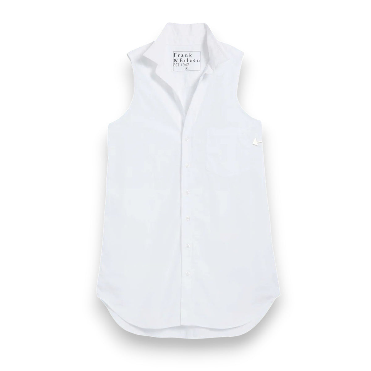 A woman smiles outdoors on a tiled patio, hands in pockets, wearing the FRANK &amp; EILEEN Finley Sleeveless Layering Button Up Shirt in Washed Linen White. Wicker chairs, a flower-topped table, and greenery complete the scene.