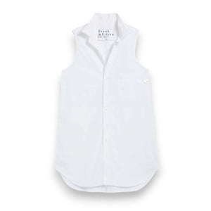 A woman smiles outdoors on a tiled patio, hands in pockets, wearing the FRANK & EILEEN Finley Sleeveless Layering Button Up Shirt in Washed Linen White. Wicker chairs, a flower-topped table, and greenery complete the scene.