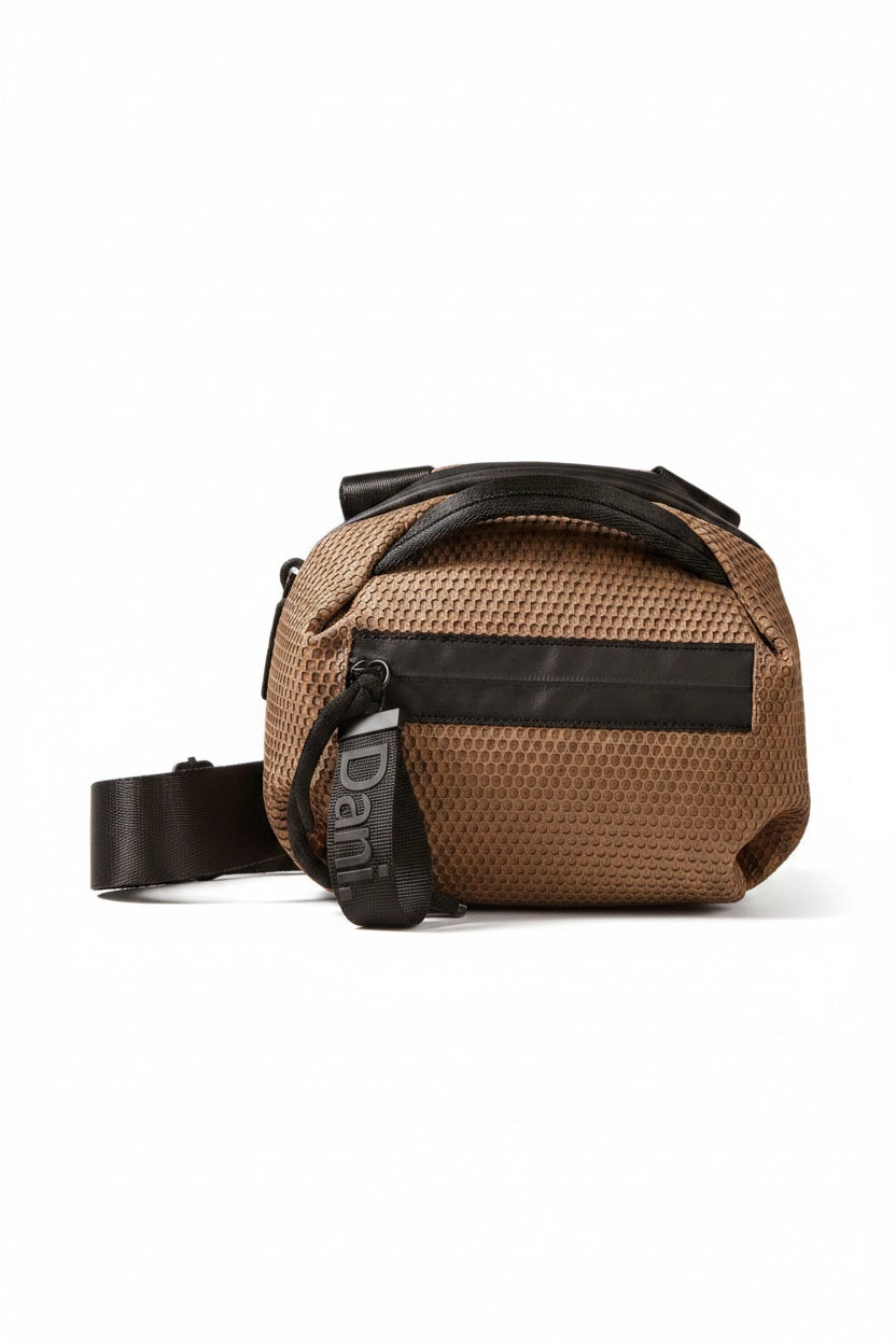 The DANIELLA LEHAVI BONNIE MINI TRAVEL CROSSBODY, a small brown vegan bag with black accents and strap, is displayed on a white pedestal against a plain beige background.
