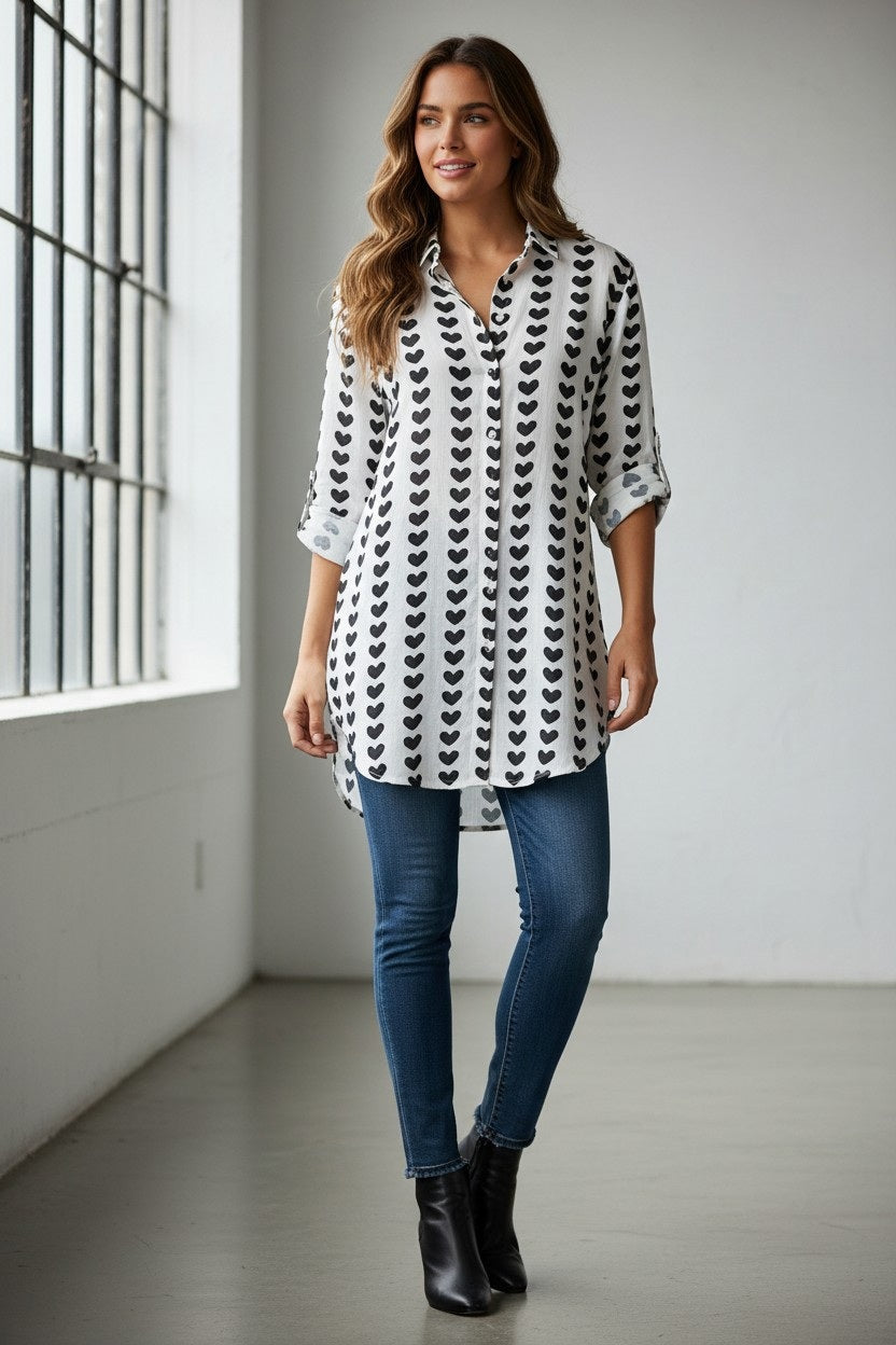A woman stands indoors wearing the EMILY PHILLIPS - THE BLACK HEARTS MINI DRESS/LONG SHIRT, featuring a straight fit and a white button-up design with a black heart pattern. She styles it with blue skinny jeans and black ankle boots. Her long, wavy hair frames her face as she smiles slightly, with one hand by her side.