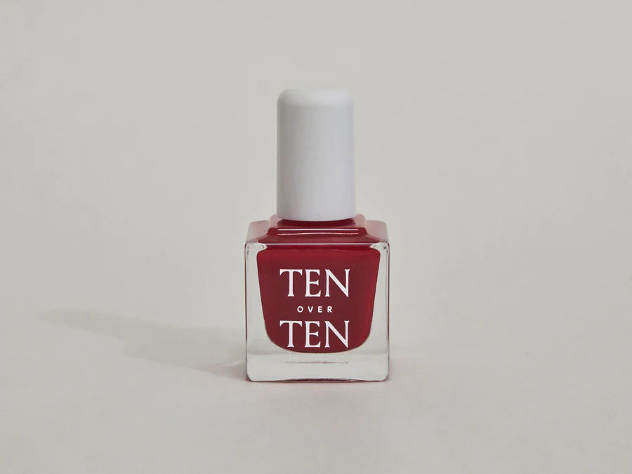 A glass bottle of TENOVERTEN - NAIL POLISH from the brand TEN OVER TEN, featuring a white cap and a dusty pink nail color. The front label displays "TEN OVER TEN" in white uppercase letters. This vegan, eight-free formula ensures both beauty and peace of mind. The background is plain and light-colored.