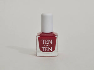 A glass bottle of TENOVERTEN - NAIL POLISH from the brand TEN OVER TEN, featuring a white cap and a dusty pink nail color. The front label displays "TEN OVER TEN" in white uppercase letters. This vegan, eight-free formula ensures both beauty and peace of mind. The background is plain and light-colored.