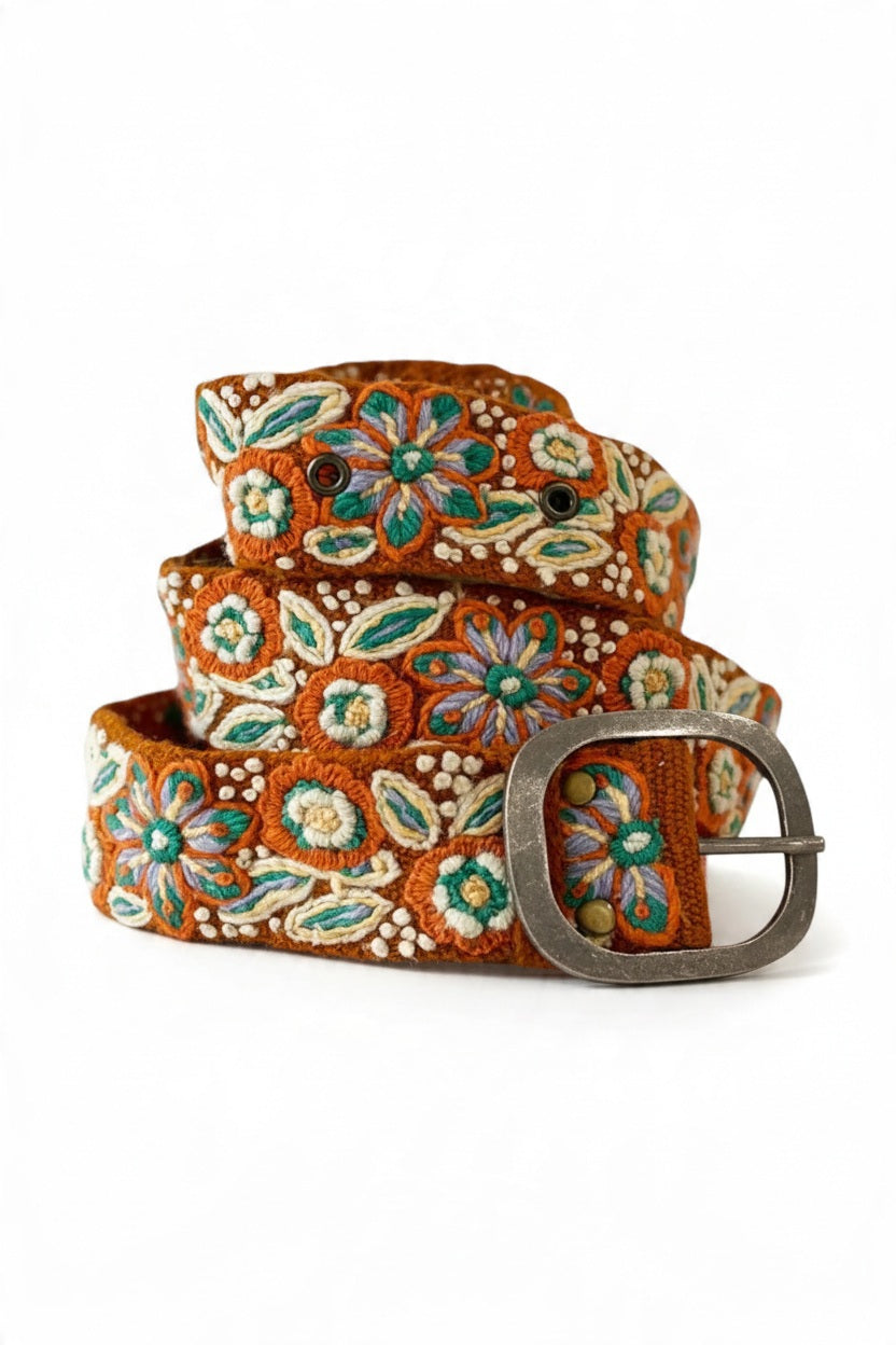 JENNY KRAUSS - GROUND COVER FLORAL EMBROIDERED WOOL BELT coiled on a surface, featuring intricate floral embroidery in green, white, and purple thread with a metallic buckle.