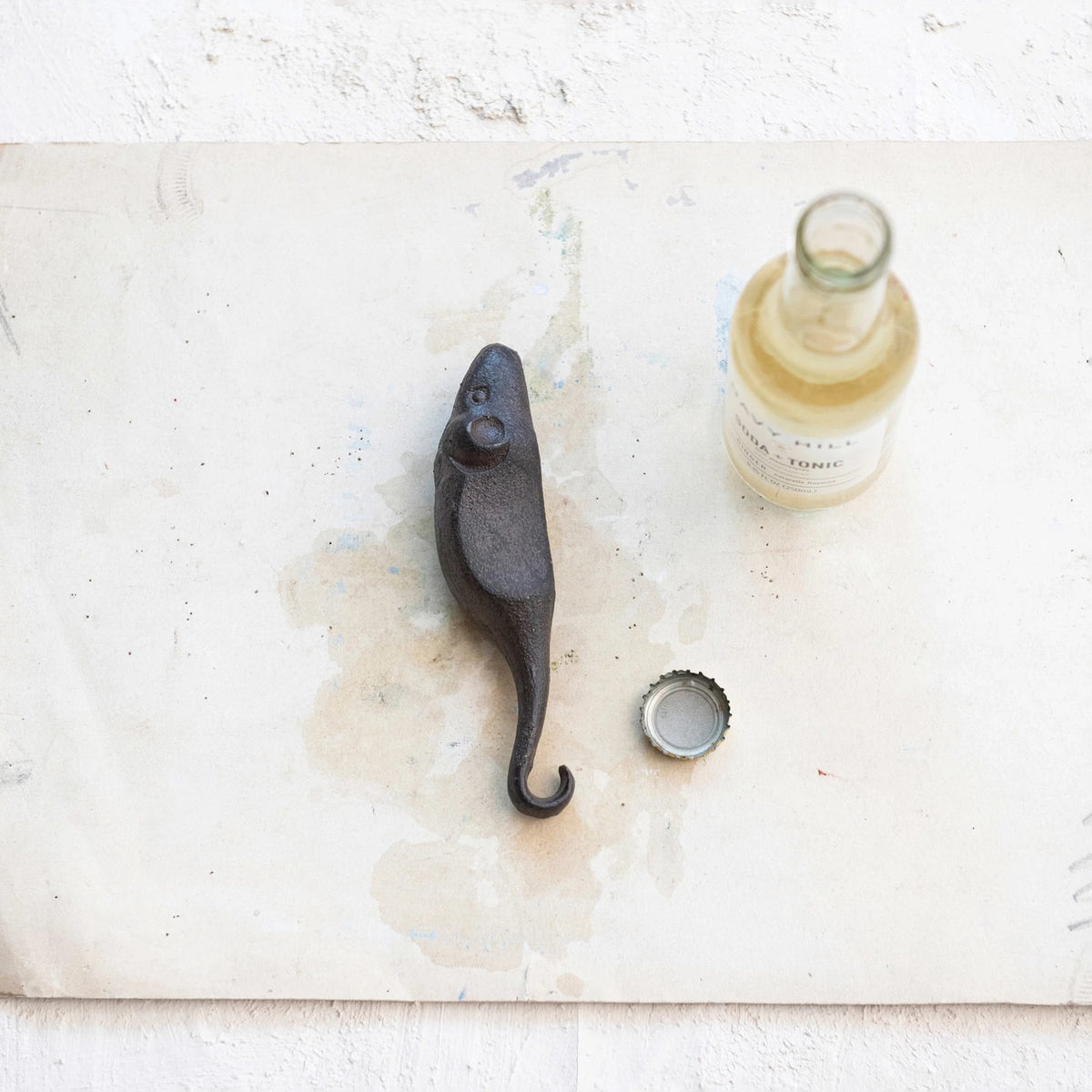 A compact, vintage-style cast iron bottle opener shaped like a mouse from Creative Coop rests on a white, slightly stained surface beside a half-full glass bottle labeled &quot;Tonic Water&quot; and its cap.