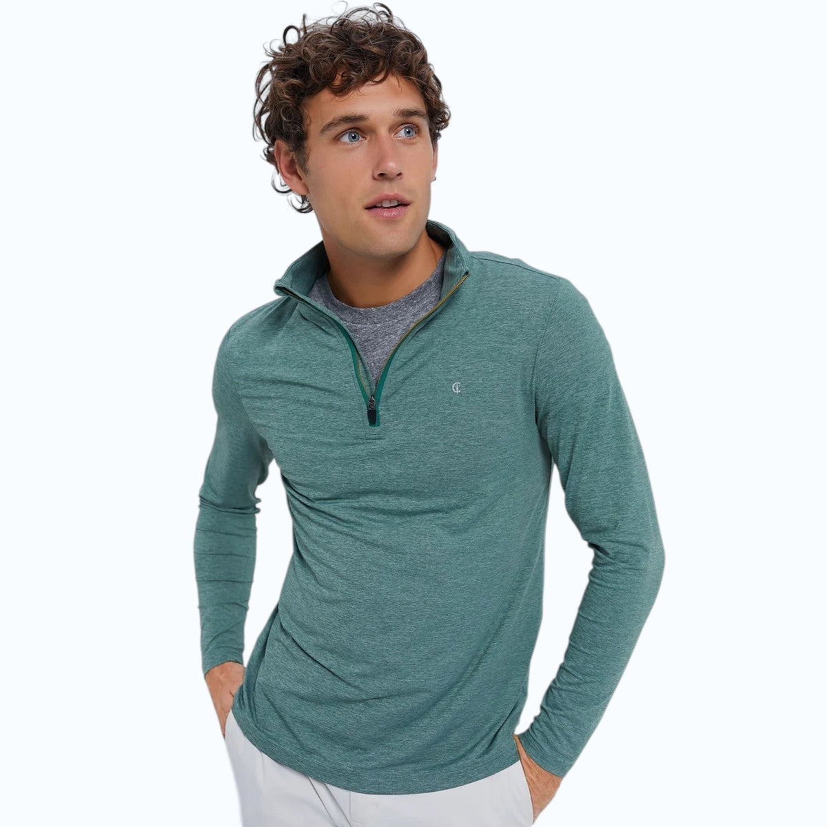 A man smiles against a plain light background, wearing the CRIQUET Feather Performance Knit Quarter Zip Pullover in Forest Heather by CRIQUET, paired with white pants and gray sneakers. His hands are in his pockets and he looks relaxed and cheerful.
