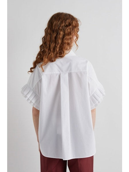 A person with long, wavy hair and sunglasses models the MEIMEIJ - WHITE COTTON SHIRT, featuring ruffled sleeves, paired with maroon pants, standing in profile against a plain white background.