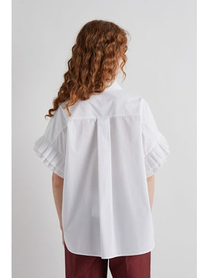 A person with long, wavy hair and sunglasses models the MEIMEIJ - WHITE COTTON SHIRT, featuring ruffled sleeves, paired with maroon pants, standing in profile against a plain white background.