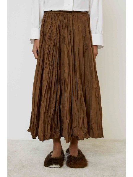 A person stands sideways on a light carpeted floor against a plain background, wearing a white long-sleeve shirt, the MEIMEIJ CRINKLE SKIRT in brown, and dark brown fuzzy slippers.