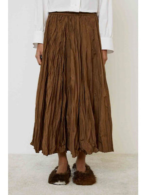 A person stands sideways on a light carpeted floor against a plain background, wearing a white long-sleeve shirt, the MEIMEIJ CRINKLE SKIRT in brown, and dark brown fuzzy slippers.