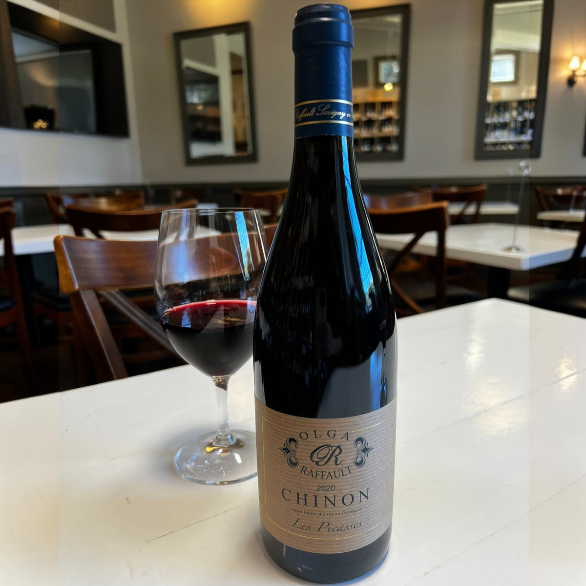 A bottle of OLGA RAFFAULT ‘LES PICASSES’ CHINON 2020 red wine from WINE: HAW RIVER WINE MAN, featuring a beige label and a dark blue foil cap, crafted from Cabernet Franc grapes and photographed against a white background.