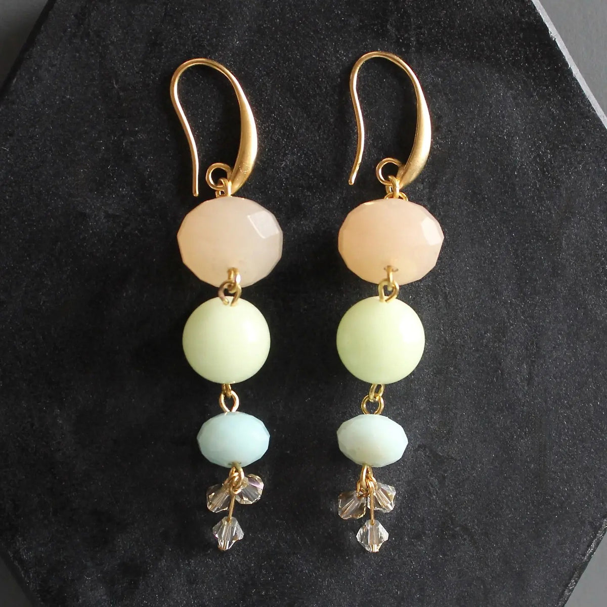 The DAVID AUBREY - JADE AND GLASS EARRINGS by DAVID AUBREY feature three vertically stacked pastel beads in peach, pale yellow, and mint green hues on gold-plated ear wires, finished with delicate crystal flower charms. These jade and glass earrings are showcased against a dark textured background.