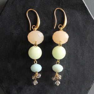 The DAVID AUBREY - JADE AND GLASS EARRINGS by DAVID AUBREY feature three vertically stacked pastel beads in peach, pale yellow, and mint green hues on gold-plated ear wires, finished with delicate crystal flower charms. These jade and glass earrings are showcased against a dark textured background.