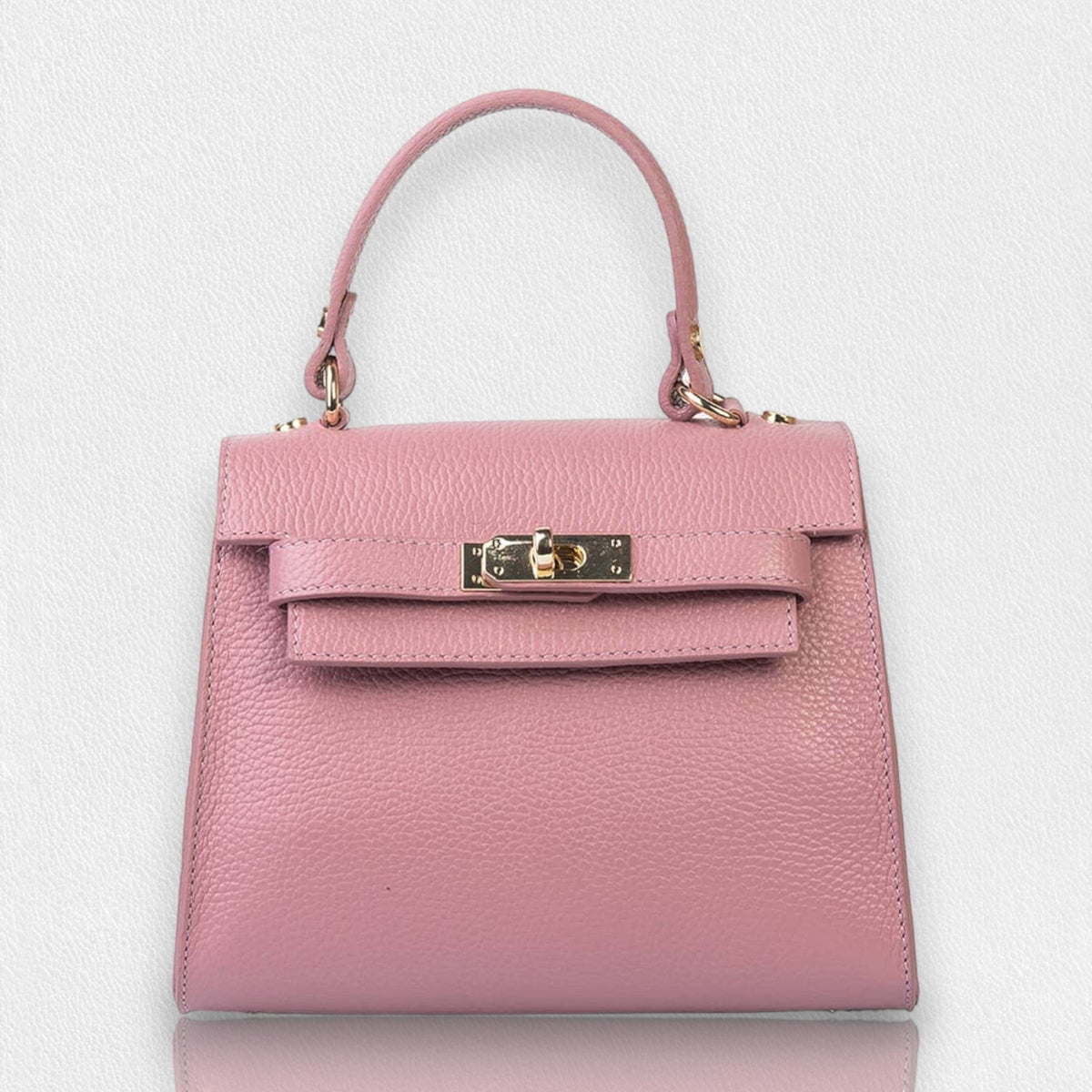 The LE BORSETTE LEATHER SATCHEL BAG is a small, structured satchel-shaped handbag in light pink, featuring a gold clasp at the front. It boasts a single top handle, an adjustable shoulder strap, and two gold-tone hardware pieces on either side of the handle. The textured leather adds to its luxurious and elegant appearance against the plain white background.