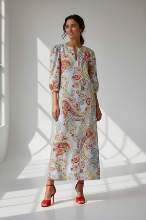 A woman with long blonde hair is seen from the back, wearing the MIRTO 1956 Paisley Print Long Sleeve Dress; its vibrant pattern and puffy sleeves flow to ankle length, revealing bright red heeled shoes against a white background.