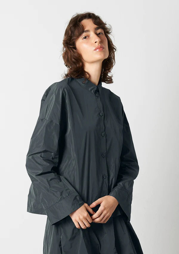 A person with shoulder-length wavy hair is wearing the KATHARINA HOVMAN Kimono Blouse from the brand KATHARINA HOVMAN. The blouse is long-sleeved, button-up, and dark gray. They are standing against a plain light gray background, gazing slightly upward with a calm expression while lightly clasping their hands in front.