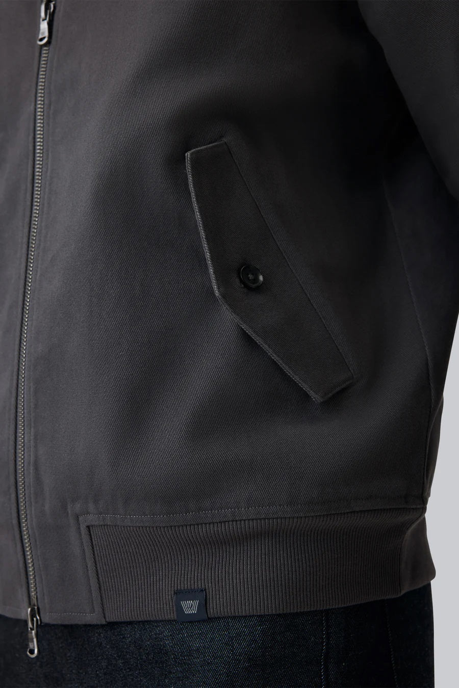 Close-up of a person wearing the MACK WELDON Twill Harrington Jacket in Shroom, featuring a zip front and buttoned flap pocket. Their arm is relaxed by their side and dark denim jeans are partially visible.