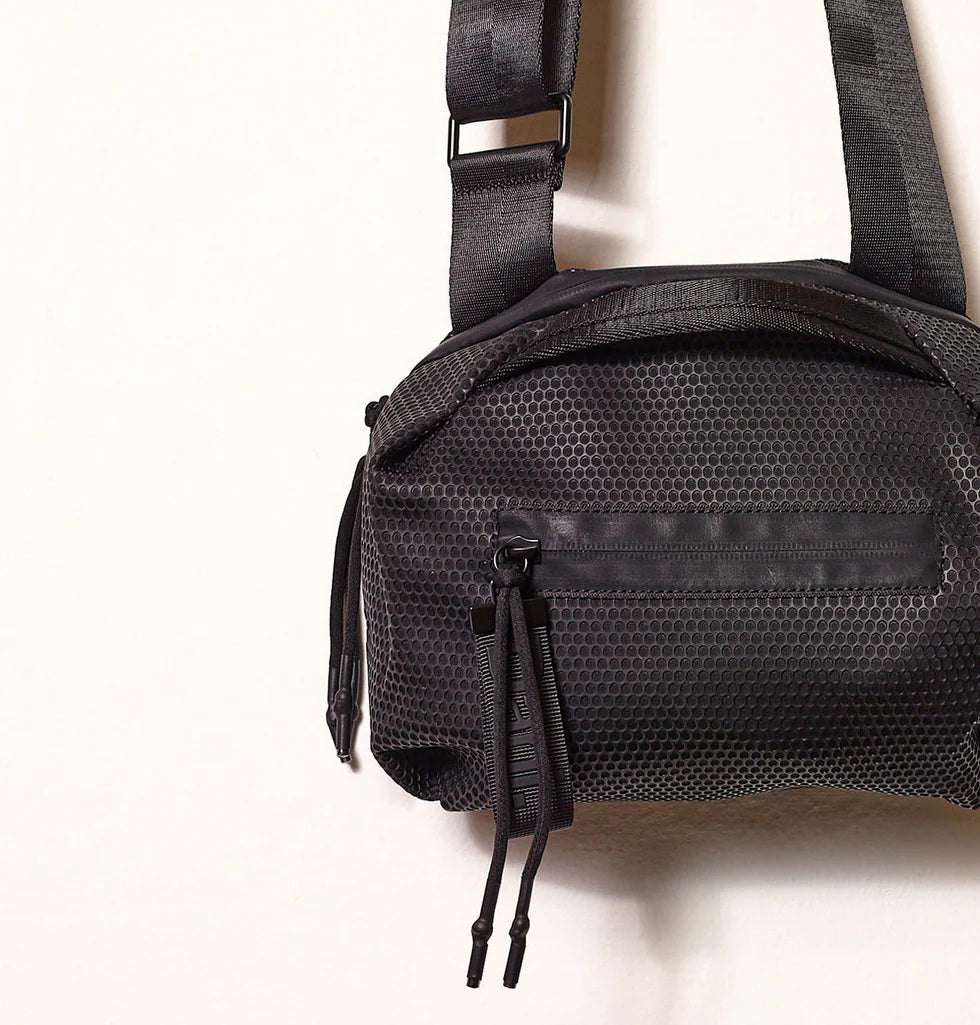 The DANIELLA LEHAVI - BONNIE TRAVEL CROSSBODY features a hexagonal textured pattern, front zipper pocket with two pulls, and an adjustable strap. Crafted from eco-friendly materials, it&#39;s shown against a plain light background.