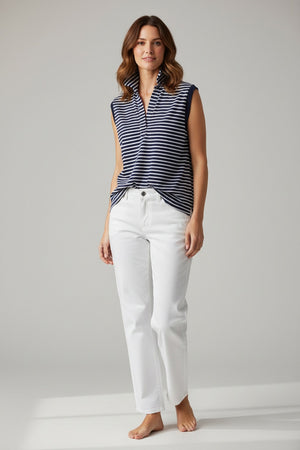A woman with short brown hair stands outdoors against a backdrop of blurred greenery, wearing the FRANK & EILEEN OLIVIA Popover Henley Tank in Vintage Jersey Yacht Stripe—a sleeveless navy and white striped shirt with a popped collar.