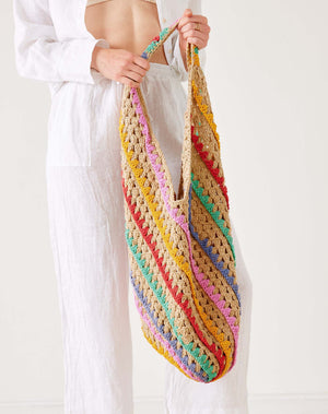 A person in white holds the MERSEA - CARLY CROCHET BOHO BAG by MERSEA, featuring large, colorful diagonal stripes of yellow, pink, red, green, and purple against a plain light background.