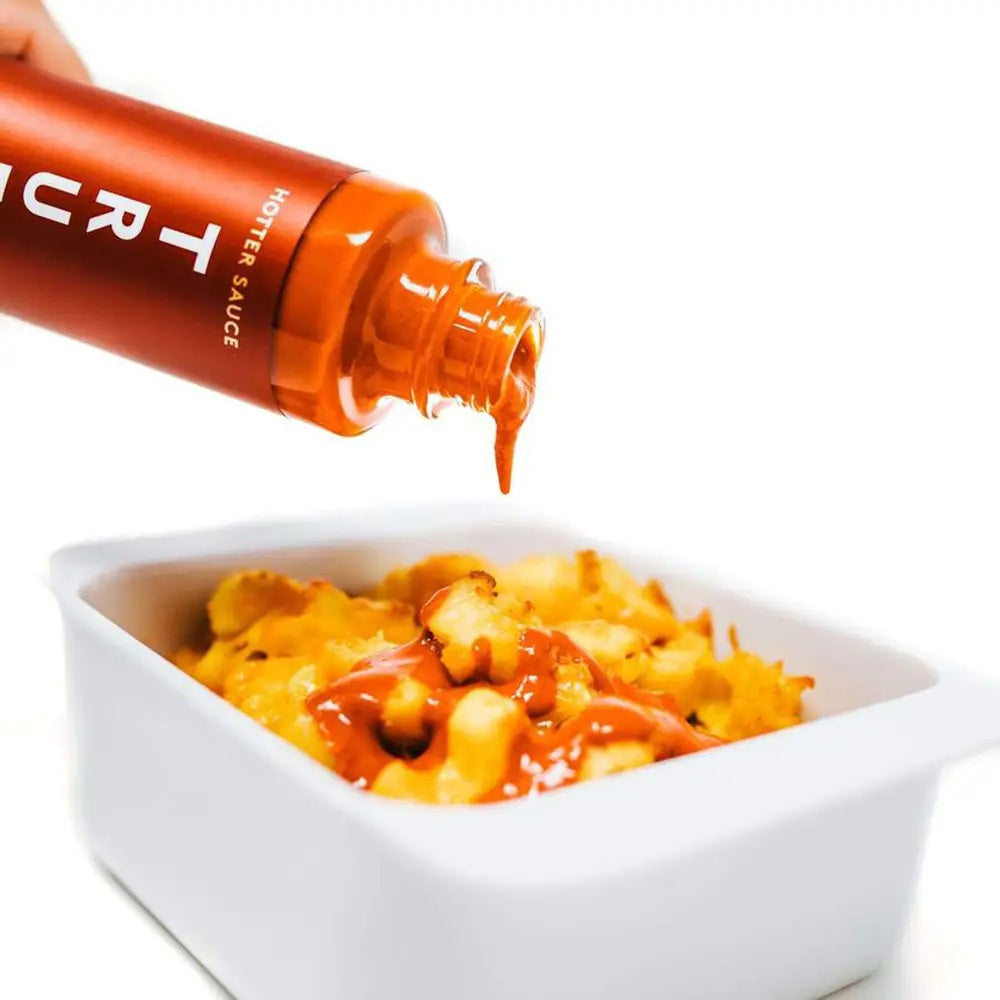 A bottle of TRUFF - HABANERO HOT SAUCE by TRUFF HOT SAUCE is being poured over a dish of macaroni and cheese in a white rectangular bowl, set against a brightly lit, white background.