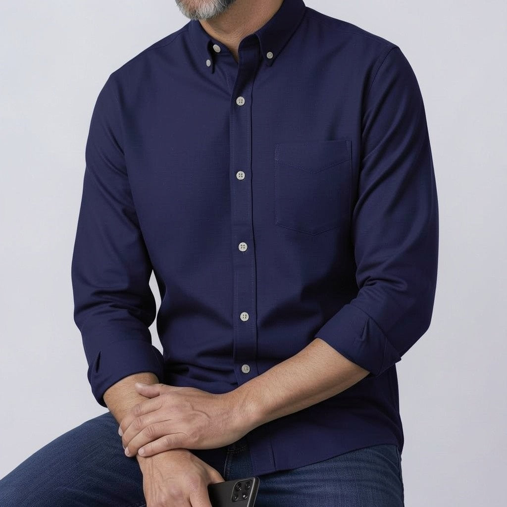 A man with short dark hair and a beard stands sideways with arms crossed, wearing the FRANK & EILEEN LUKE Men's Button Up Shirt in Famous Denim Washed Navy, blue jeans, and white sneakers. He is indoors by a light wall.