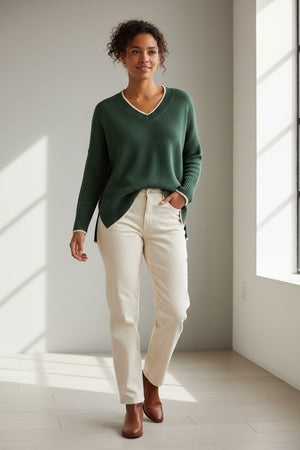 A person wearing the MERSEA Montauk Colorblock V Neck Sweater in Eden, paired with off-white pants and brown ankle boots, stands in a sunlit room with white walls and light wood floors.