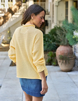 A woman with long brown hair smiles softly outdoors, wearing the FRANK & EILEEN Monterey Pointelle Rolled Funnel Neck Sweater in lemon yellow and a denim skirt. Greenery and a building with stairs appear in the background.