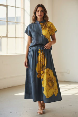 A woman stands in a sunlit room, smiling with her hands in her pockets, wearing navy trousers, open-toe shoes, and the MIRTO 1956 Short Sleeve Cotton Shirt by MIRTO 1956, featuring gray and yellow floral patterns.