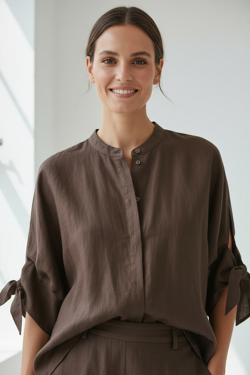A person with straight blonde hair wears the MEIMEIJ Linen Top with Tie Sleeve in Moka—a loose-fitting, dark brown linen blouse featuring short sleeves and side ties—paired with matching pants. Made in Italy by MEIMEIJ, this effortlessly chic summer top is styled against a plain light background.