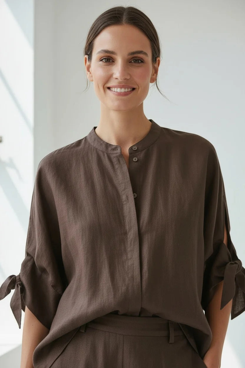 A smiling woman wearing a dark brown, long-sleeved, button-down blouse with tie-detail cuffs.