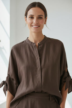 A person with straight blonde hair wears the MEIMEIJ Linen Top with Tie Sleeve in Moka—a loose-fitting, dark brown linen blouse featuring short sleeves and side ties—paired with matching pants. Made in Italy by MEIMEIJ, this effortlessly chic summer top is styled against a plain light background.