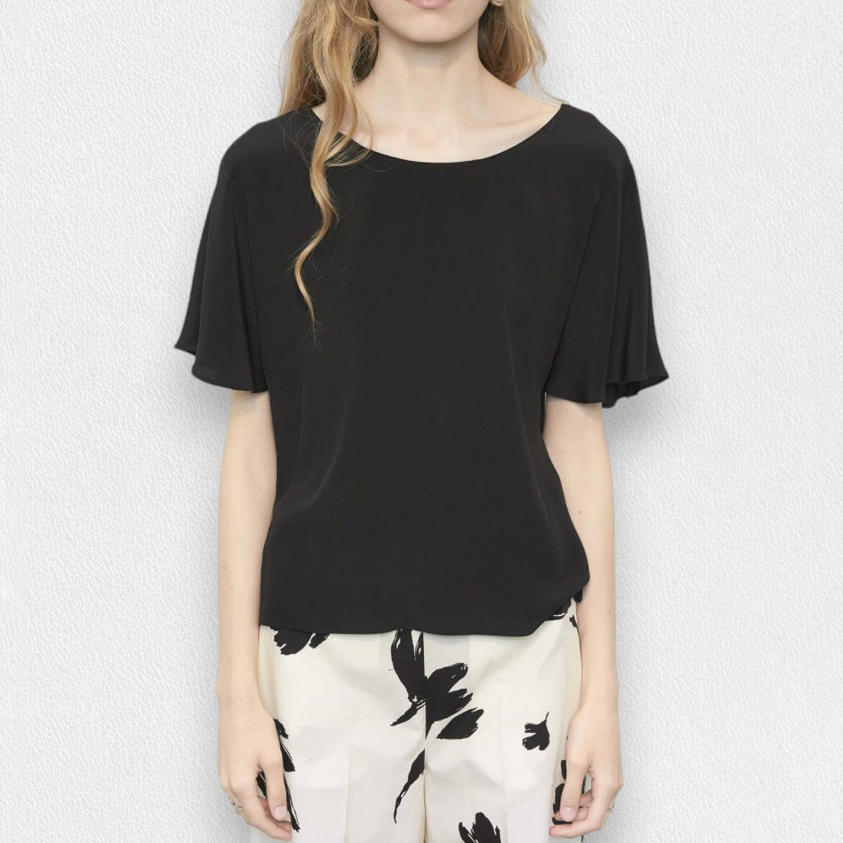 A woman with long blonde hair is wearing a black, short-sleeved MEIMEIJ - Flutter Sleeve Blouse and white pants with a black floral pattern. She is standing against a plain, light-colored background. Her face is not visible in the image.