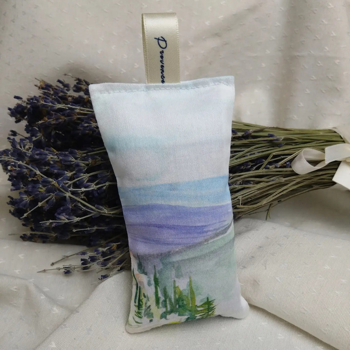 A L&#39;AUGUSTE PROVENCE French Lavender Sachet from Provence, adorned with a cream &quot;L’AUGUSTE&quot; ribbon and painted with a hillside village, hangs before dried lavender tied with white ribbon.