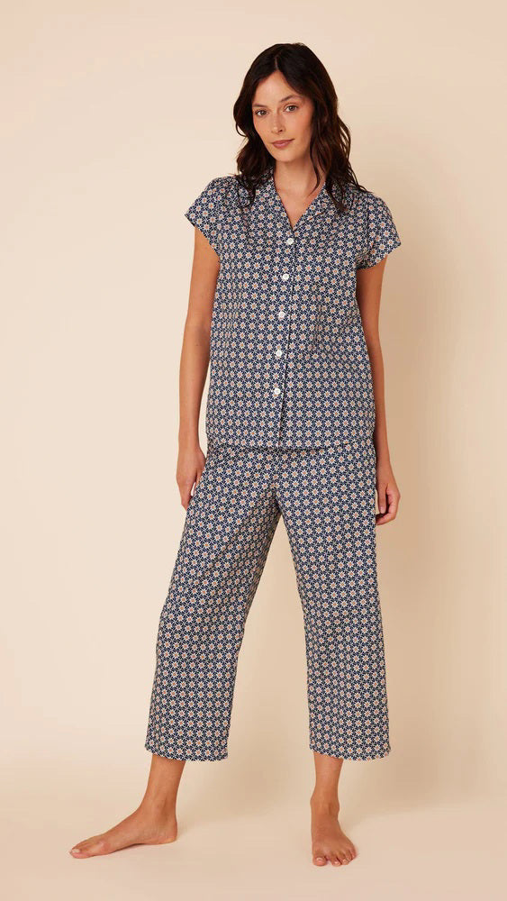 A woman stands barefoot against a plain beige background, wearing the CAT&#39;S PAJAMAS - NEWPORT DAISY LUXE PIMA COTTON CAPRIS PAJAMA from CAT&#39;S PAJAMAS. The short-sleeved button-up top and matching loose-fitting capri pants feature a small, intricate pattern in shades of blue and white, crafted from 100% Pima cotton.