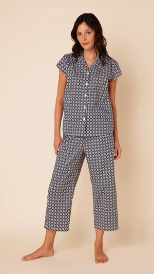 A woman stands barefoot against a plain beige background, wearing the CAT'S PAJAMAS - NEWPORT DAISY LUXE PIMA COTTON CAPRIS PAJAMA from CAT'S PAJAMAS. The short-sleeved button-up top and matching loose-fitting capri pants feature a small, intricate pattern in shades of blue and white, crafted from 100% Pima cotton.