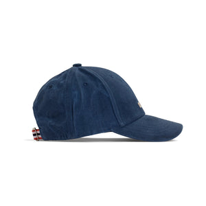 The AMUNDSEN SPORTS - WAXED COTTON CAP in navy blue is shown from the side against a plain white background, featuring a curved brim and an adjustable strap with a red, white, and black checkered norgesreima design.