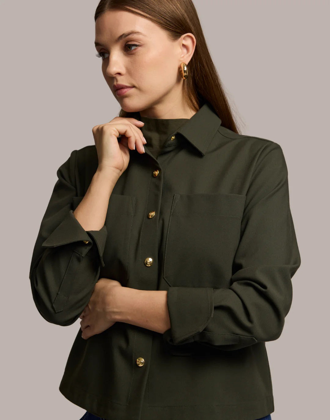 A woman with long brown hair models the HINSON WU - ASTRID LONG SLEEVE TRAVELER&#39;S TWILL SHIRT JACKET in dark green over an olive top and blue jeans, showcasing versatile HINSON WU travel style with gold hoop earrings against a neutral background.
