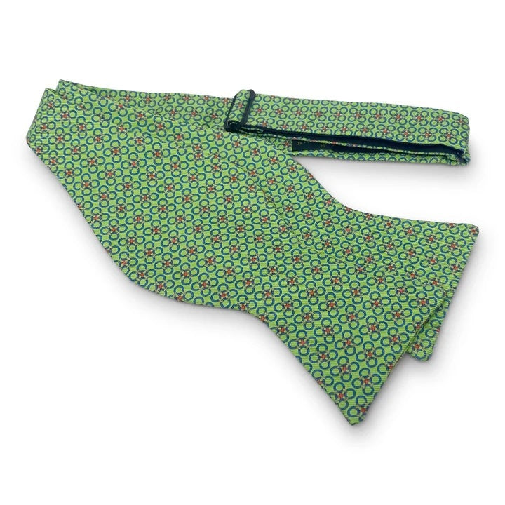 The PETER-BLAIR - CIRCLE CROSS SILK BOW TIE IN GREEN by BIRD DOG BAY features a green adjustable design adorned with a repeating pattern of blue and red rings, shown against a white background.