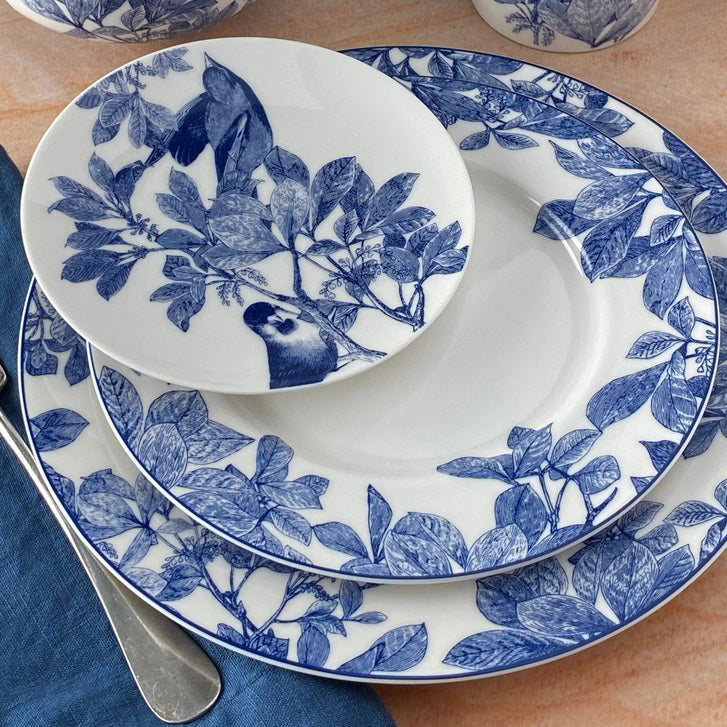 Three CASKATA ARBOR BLUE BIRDS PLATES, featuring blue and white botanical and bird motifs on premium porcelain, are stacked on a table beside a silver spoon and a folded blue napkin.