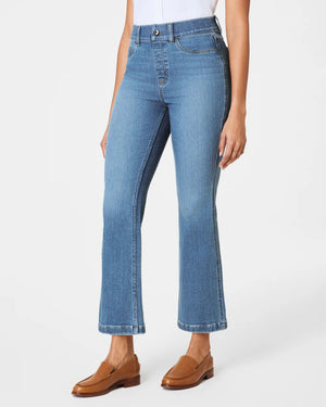 A person poses against a plain backdrop, wearing a sleeveless white blouse and SPANX Pull On Kick Flare Jeans with brown loafers. A beige jacket is draped over their shoulders, and their hair is styled in a bun.