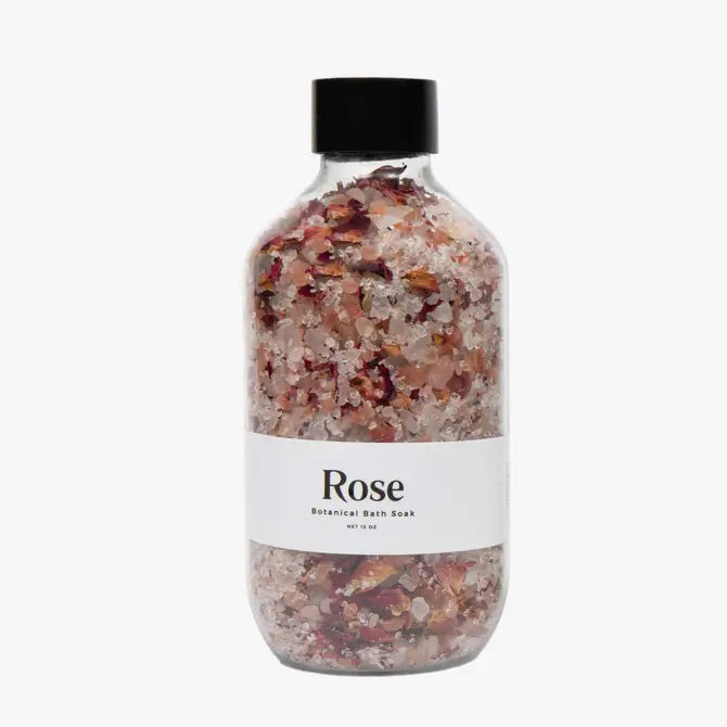 A transparent glass bottle containing a blend of pink and white salt crystals and rose petals, enriched with organic rose and lavender essences. The bottle features a white label that reads &quot;BOTANICAL BATH SOAK - ROSE&quot; from GIFTEN MARKET and is topped with a black cap. Set against a white background, it promises an indulgent bathing experience.