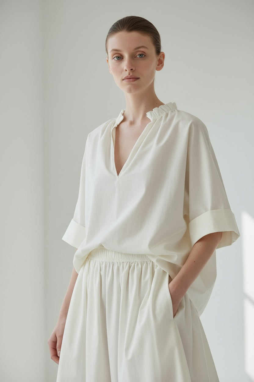 A person wearing the MEIMEIJ Gathered Neck Blouse in Off White by MEIMEIJ stands against a plain light background, looking calmly at the camera with hands in pockets.
