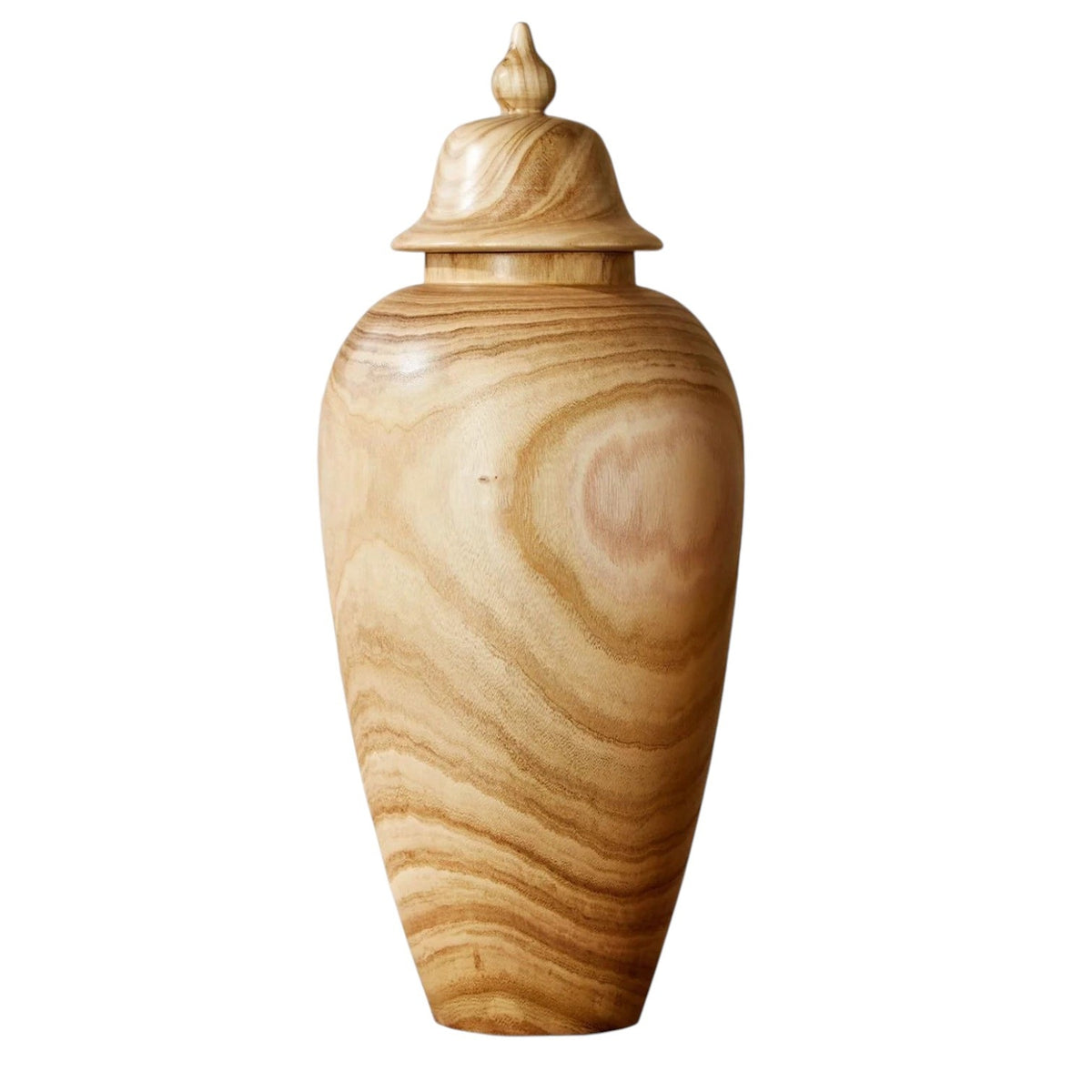 The PAULOWNIA WOOD COVERED TEMPLE JAR - LARGE by TWO'S COMPANY rests elegantly on a wooden surface before a light brick wall, its tall silhouette and swirling grain patterns enhanced by a fitted lid. Green plant leaves peeking in from the right complete this exotic home accessory, bringing warmth and character to any space.