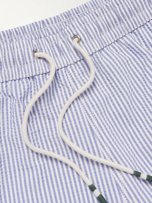 A person wears HARTFORD - SEERSUCKER SWIM TRUNKS IN INDIGO with a drawstring waist, side pockets, and a quick-drying seersucker fabric. The mid-thigh trunks are shown against a plain background, displaying only the torso and legs.