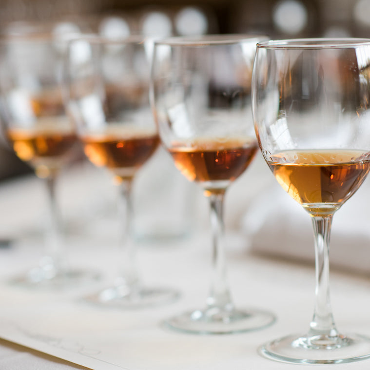 Four wine glasses partially filled with amber-colored liquid are lined up in a row on a white surface, with a blurred background.