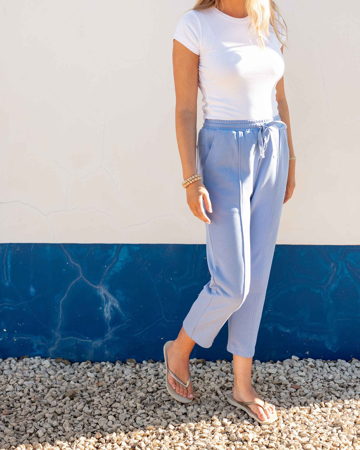 A person wears the MERSEA - FRENCHIE CIGARETTE PANT by MERSEA, paired with a white top and gray sneakers, standing on sandy beach ground with ocean waves and blue sky behind them, hands in their pockets.