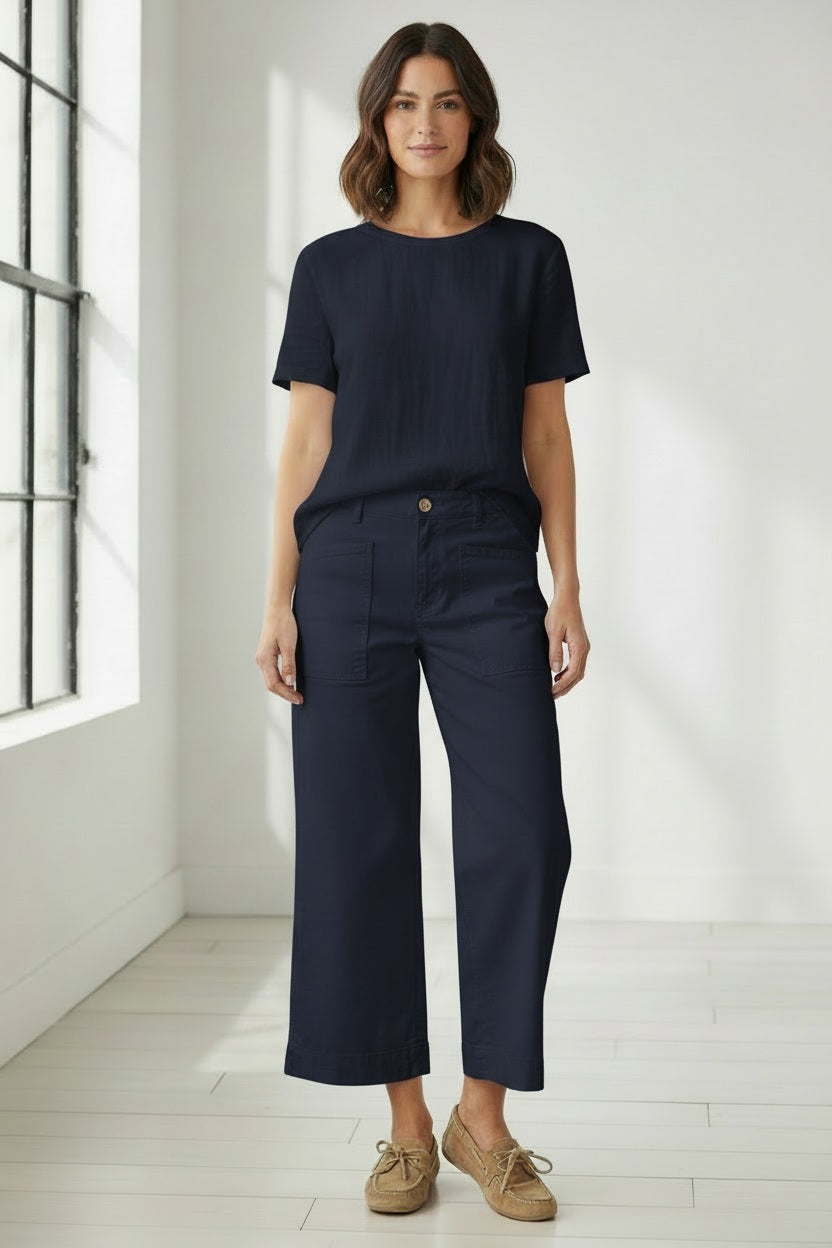 A woman stands indoors by a window, wearing a navy Italian Performance Linen short-sleeve top, FRANK & EILEEN’s LIMERICK Wide Leg Carpenter Pant in Classic Navy, and tan boat shoes. The room has white walls and light wooden floors.