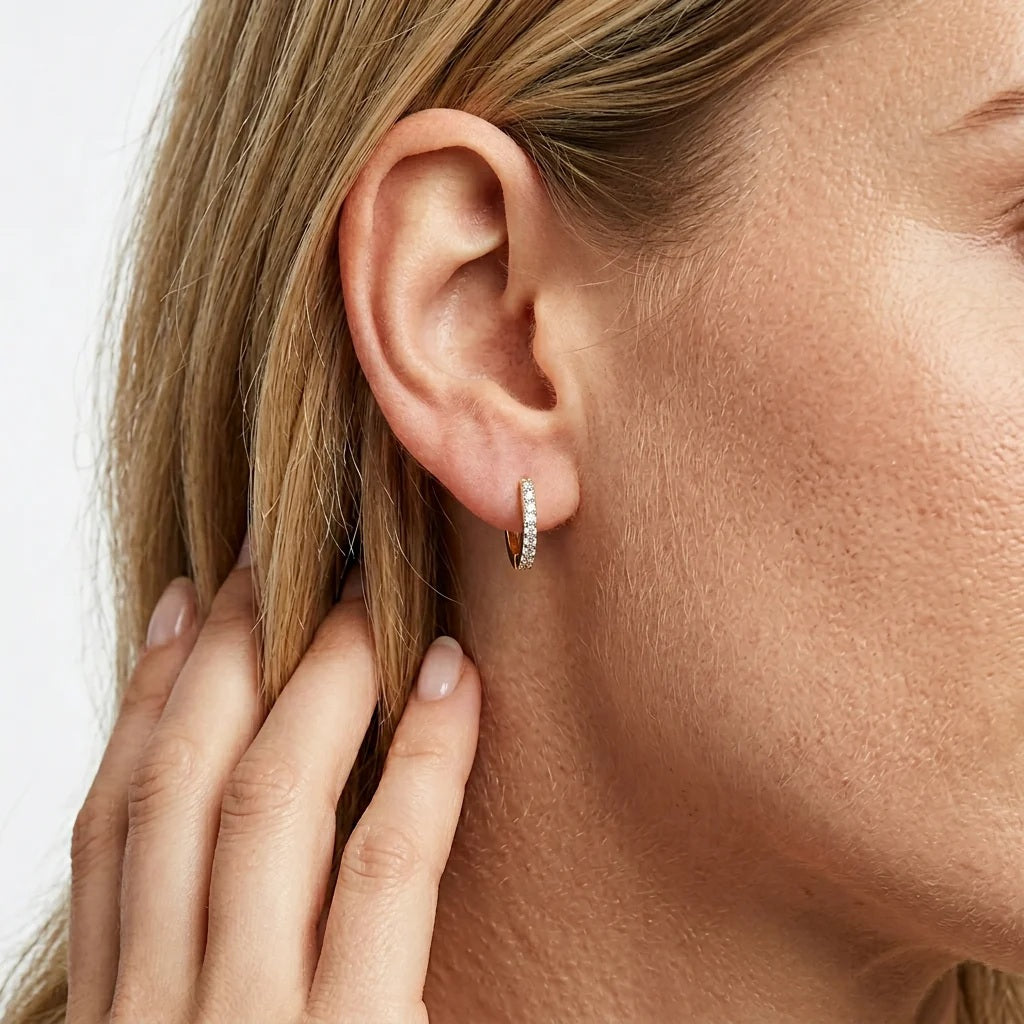 A close-up of a woman’s ear wearing the ELLA STEIN - CLASSIC PAVE DIAMOND HUGGIE EARRINGS (LARGE) in gold, adorned with natural diamonds. Her hand, featuring neatly manicured nails, gently touches her hair near her ear.