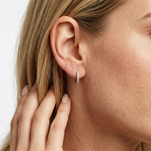 A close-up of a woman’s ear wearing the ELLA STEIN - CLASSIC PAVE DIAMOND HUGGIE EARRINGS (LARGE) in gold, adorned with natural diamonds. Her hand, featuring neatly manicured nails, gently touches her hair near her ear.