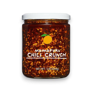 A clear jar of MOMOFUKU – CHILI CRUNCH shows chili flakes, crunchy garlic, and oil. The label features orange art and white text with the product name and 150g (5.3oz), all displayed against a plain white background.